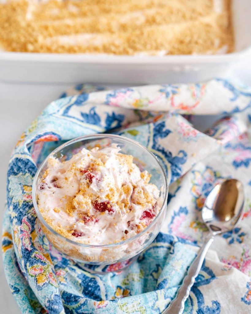 Top down view of a glass dish filled with a Strawberry Cheesecake Salad that's been garnished with graham cracker crumbs. A white baking dish filled with the salad sits in the background.