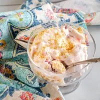 Top down view of a glass dish filled with a Strawberry Cheesecake Salad that's been garnished with graham cracker crumbs. A white baking dish filled with the salad sits in the background.