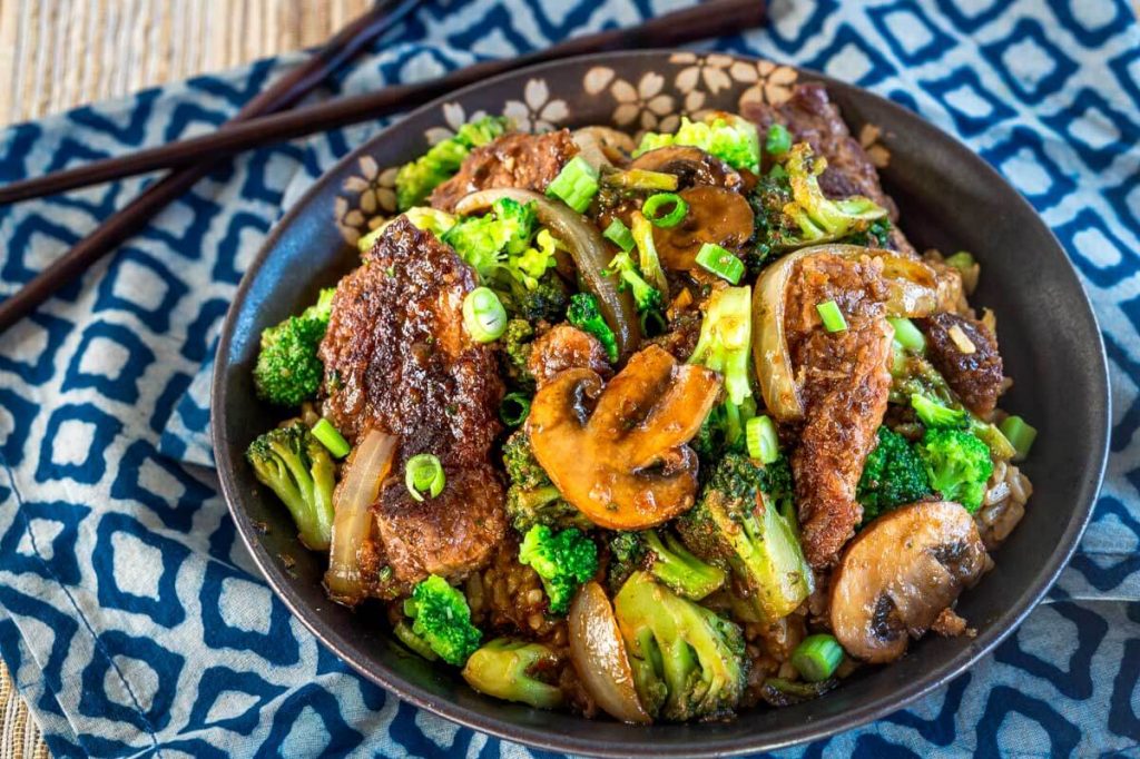 Top down view of a black bowl filled with stir fried beef, broccoli, mushrooms, and onions over a blue checked napkin. A set of chop sticks sit in the background.