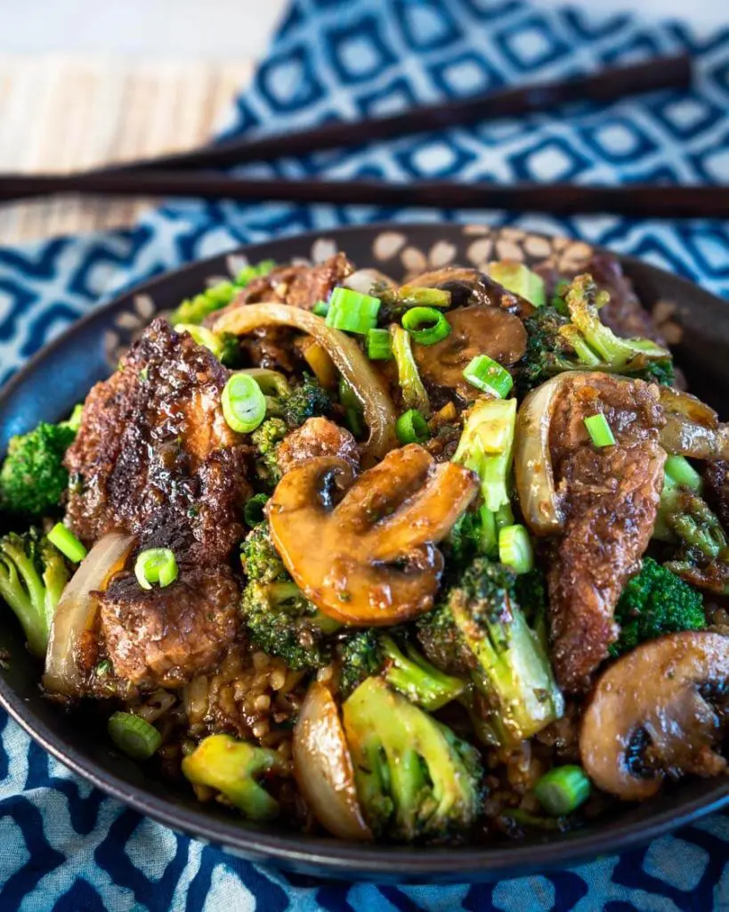Angled view of a black bowl filled with stir fried broccoli, beef, mushrooms, and onions over a blue checked napkin. A set of chop sticks sit in the background.