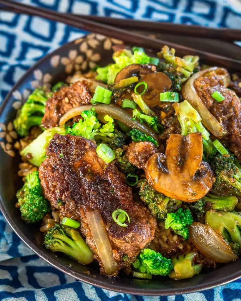 Close up top down view of a black bowl filled with Beef and Broccoli Stir Fry over a blue checked napkin. A set of chopsticks sits on the back of the bowl.