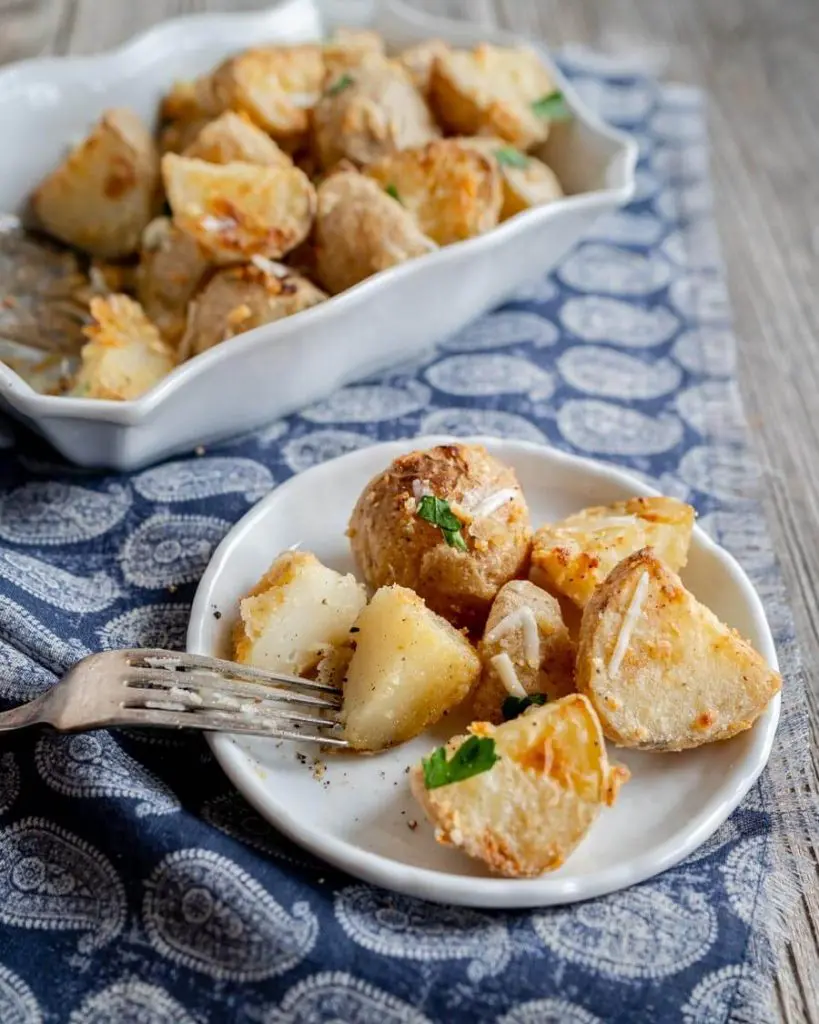 A plate of parmesan crusted roasted potatoes with one cut in half. A baking dish filled with potatoes sits in the background.