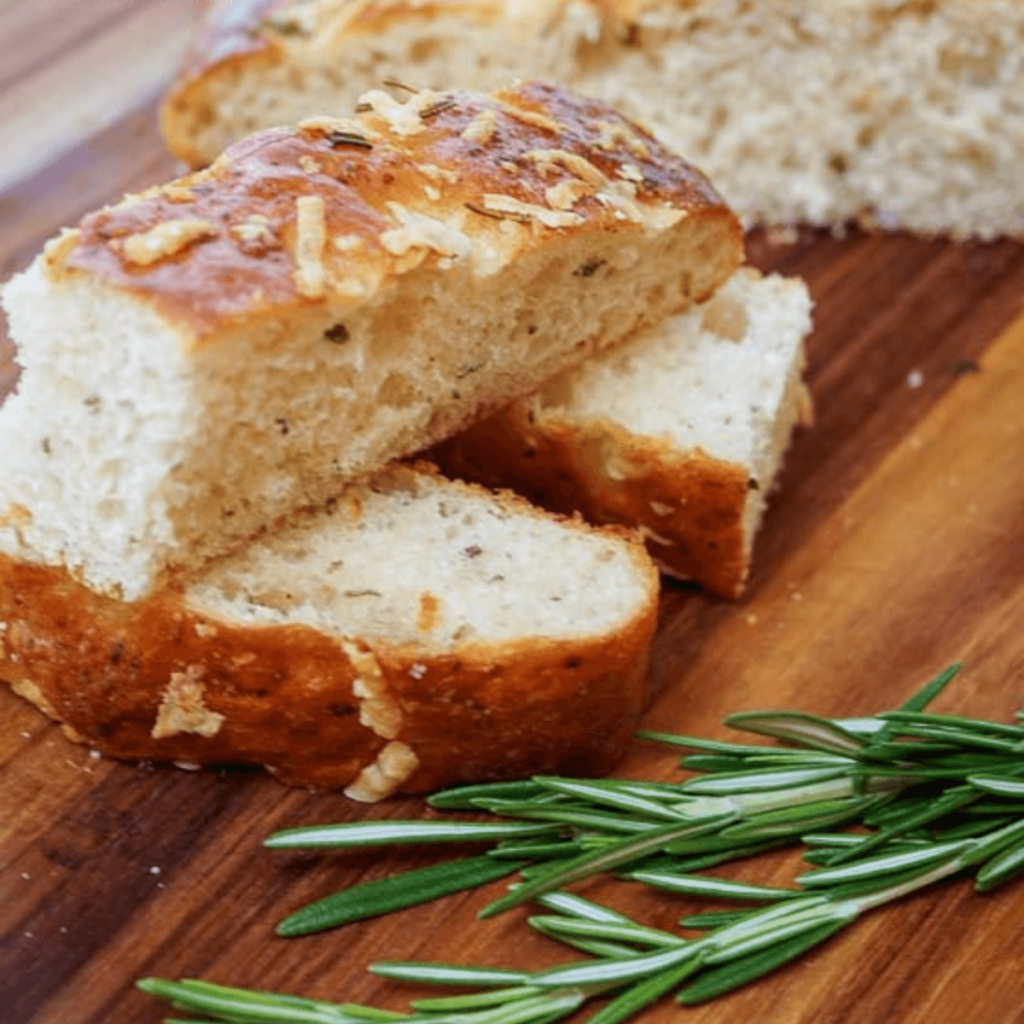 Side view of sliced Focaccia bread topped with cheese and herbs on a wooden cutting board. Fresh rosemary sits in the front.