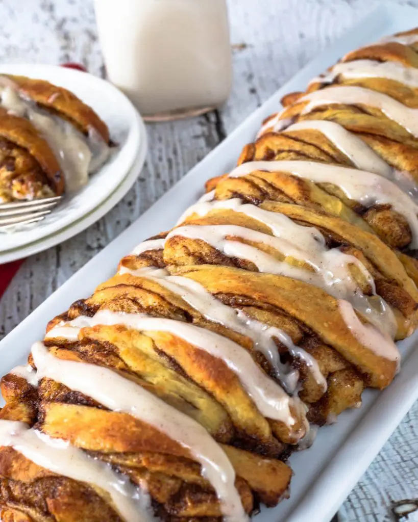 Top down view of a loaf of cinnamon pull-apart bread drizzled with a glaze. A plate of a slice of the bread sits in the background next to a bottle of milk.