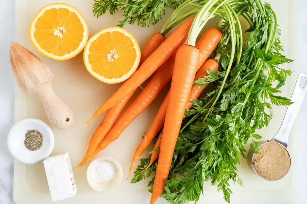 Top down view of a cutting board filled with fresh carrots, oranges, butter, brown sugar, salt and pepper.