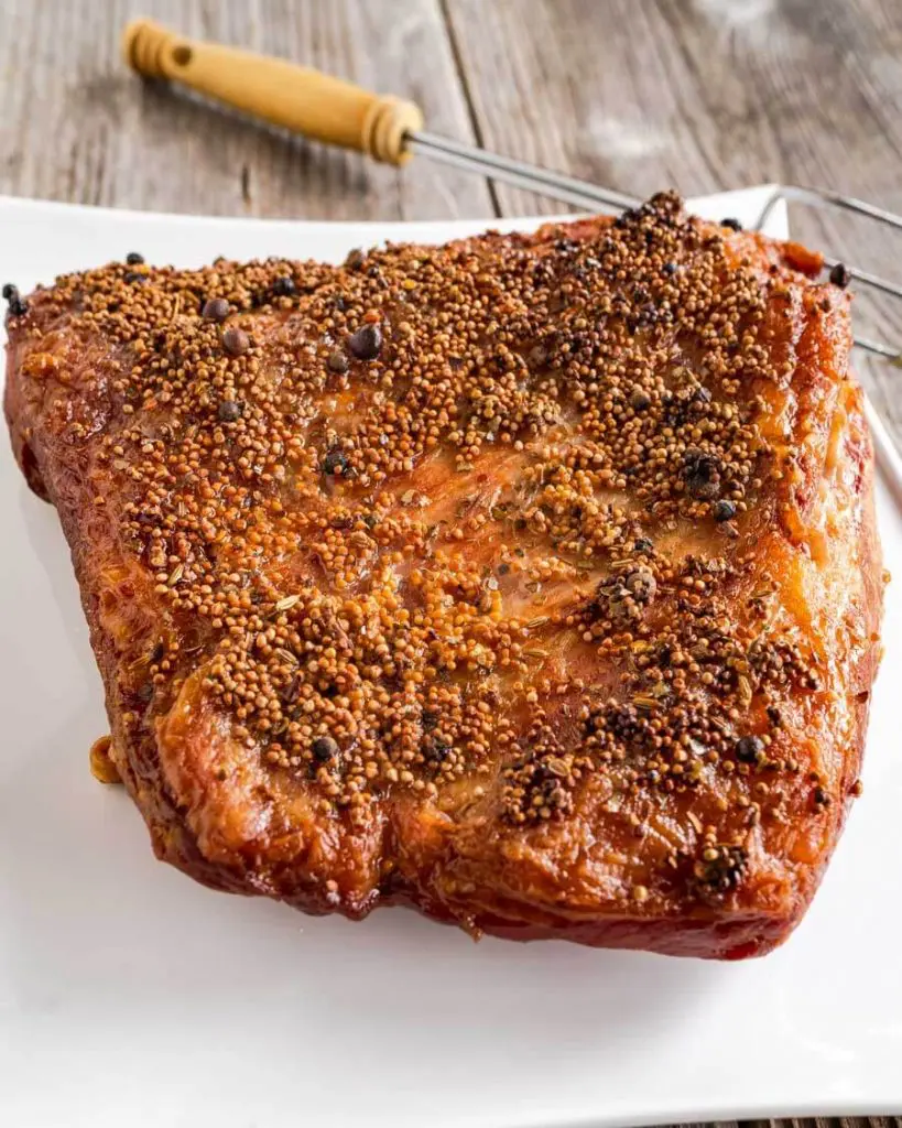 3/4 view of a baked corned beef brisket covered with spices sitting on a white platter with a serving fork in the background.