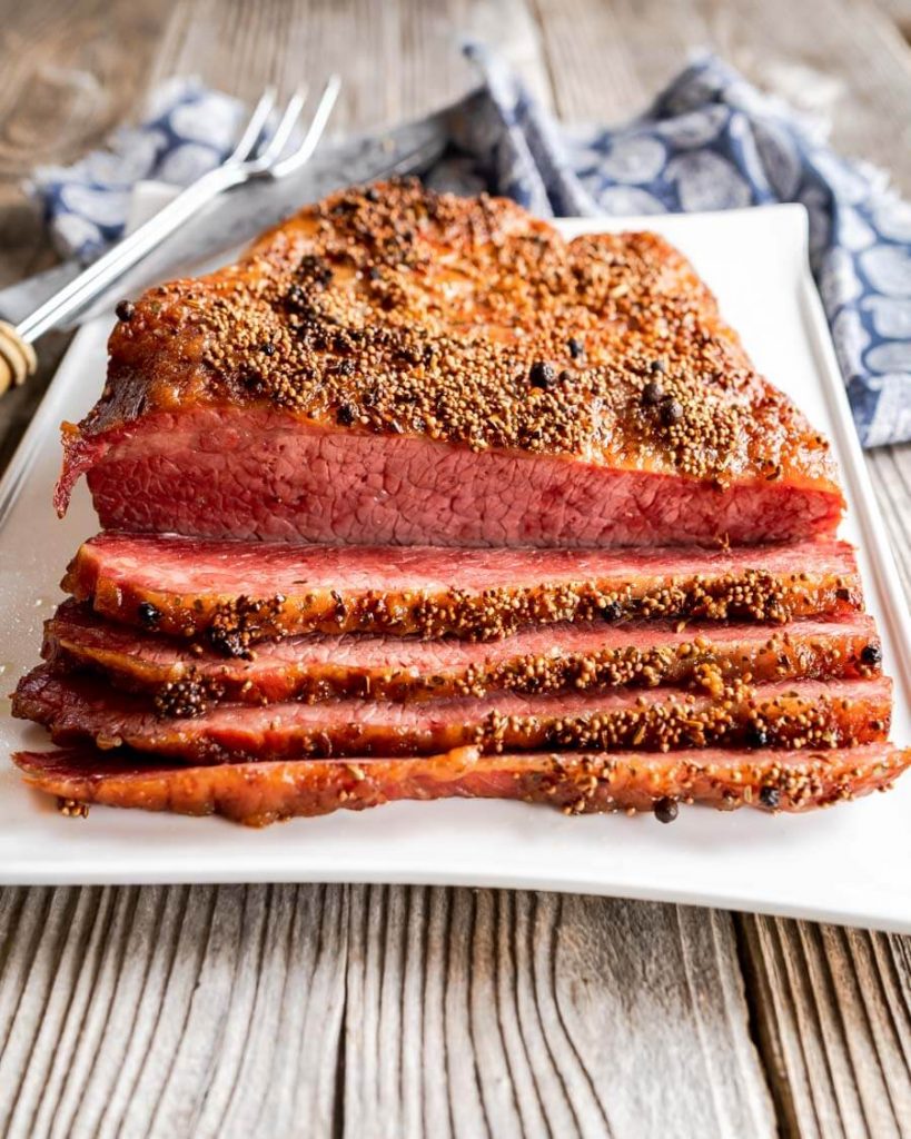 Front view of a baked corned beef brisket with 4 slices cut from the front showing the dark pink interior sitting on a white platter over a blue patterned napkin.
