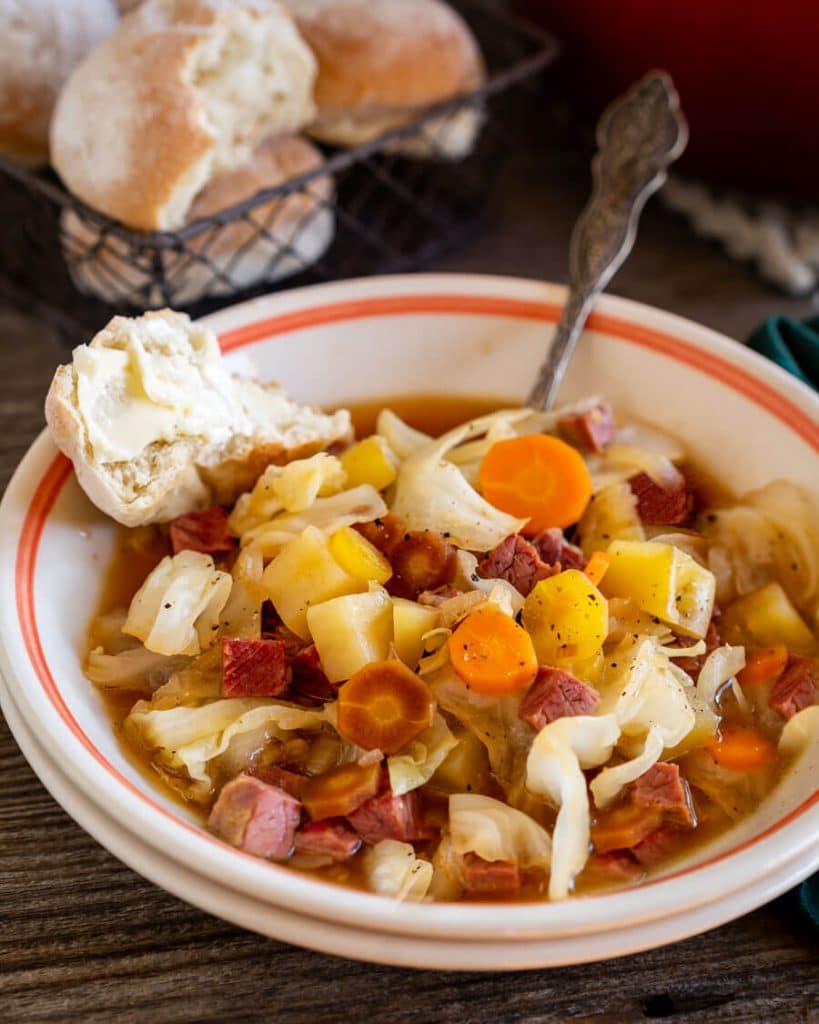 Close up view of a bowl of corned beef and cabbage soup with rolls in the background.