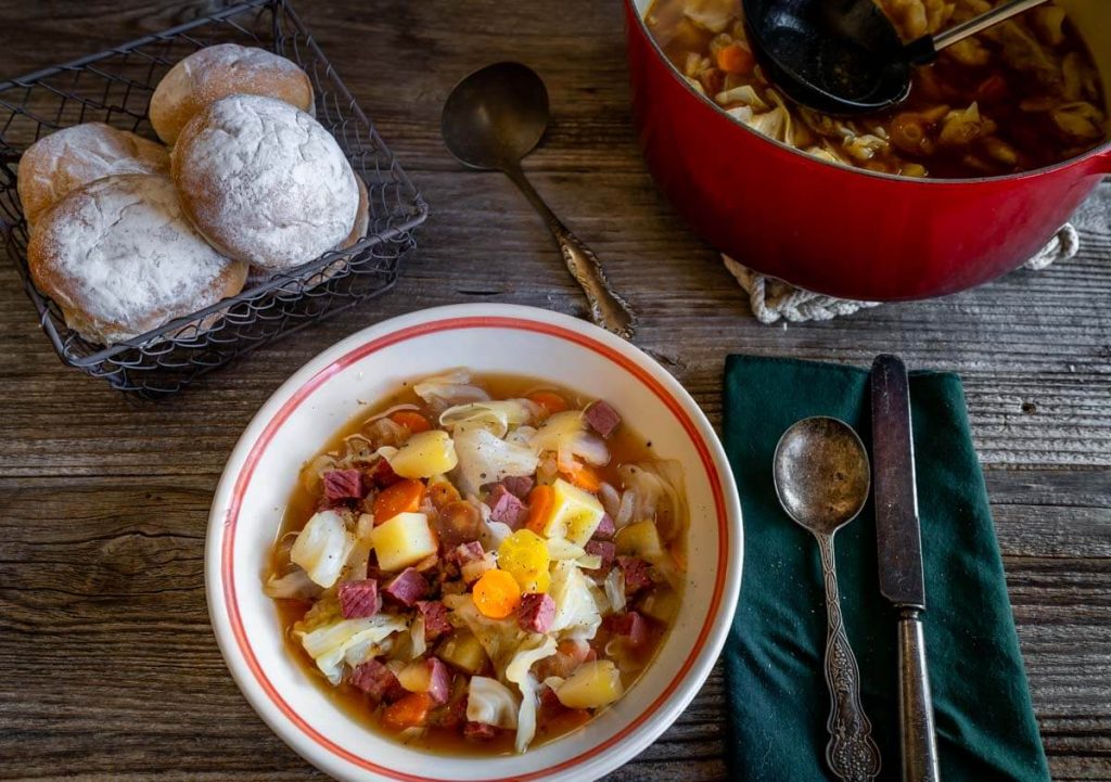 Top down photo of a bowl of corned beef and cabbage soup. A basket of rolls and a pot of soup sit in the background. A setting of silverware sit to the side of the bowl.