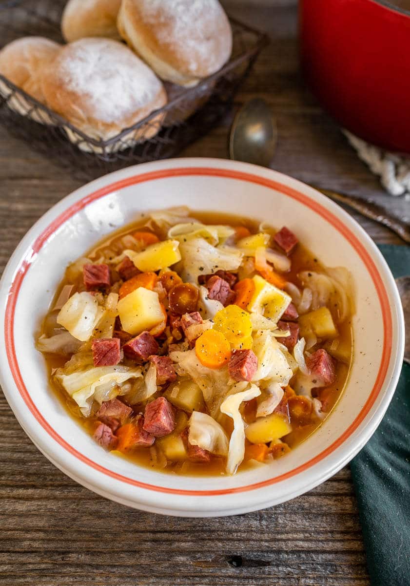 3/4 angle of a bowl of corned beef and cabbage soup with rolls in the background.