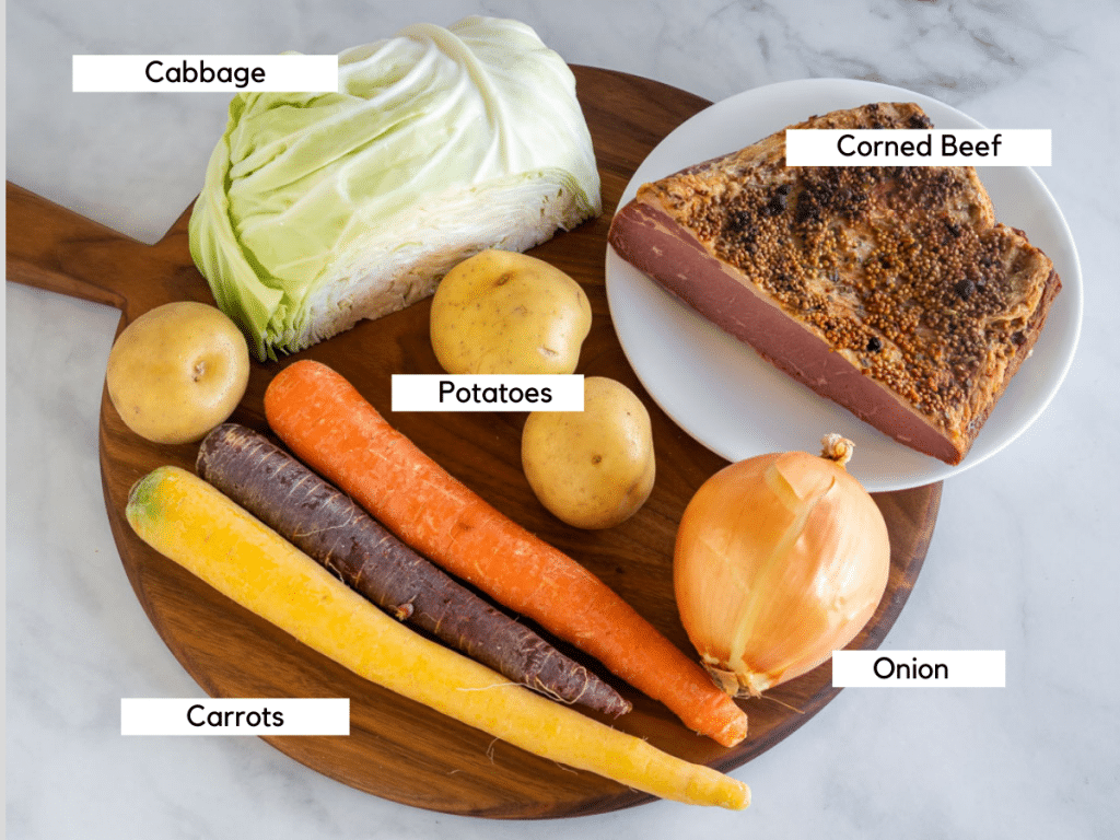 Carrots, potatoes, onion, cabbage, and corned beef brisket on a wooden cutting board.