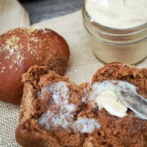 Close up shot of a brown bread roll split in half and spread with butter. A whole roll and jar of butter sit in the background.