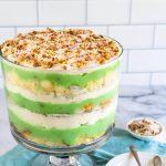 Side-view photo of a trifle bowl filled with pistachio pudding, cake, whipped cream and topped with toasted coconut with pecans.