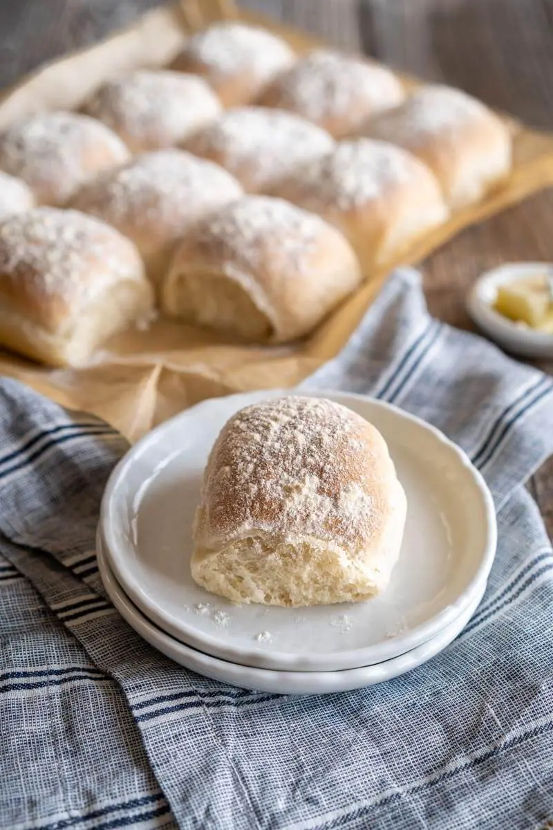 A baked roll dusted with flour sitting on a white plate with the pan of rolls sitting in the background.
