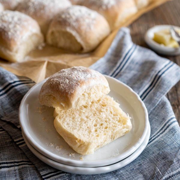 Close up of a baked bread roll cut in half showing the soft center sitting on a plate with the pan of baked rolls in the background.