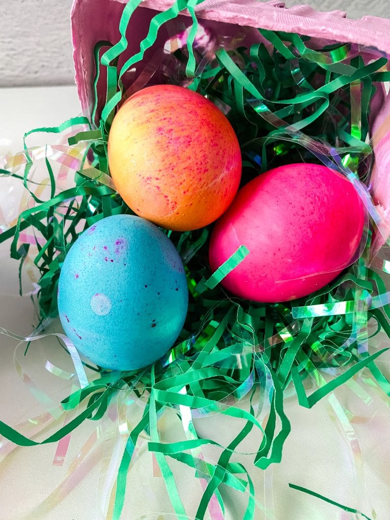 Top down photo of colored eggs in shredded green paper next to a pink basket.