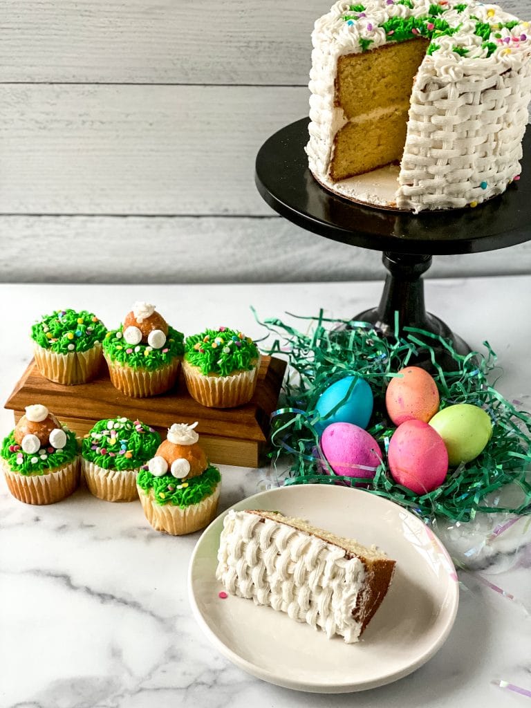 A beautiful basket weave cake on a display stand with one piece on a plate, shown with bunny butt cupcakes and colored Easter eggs.