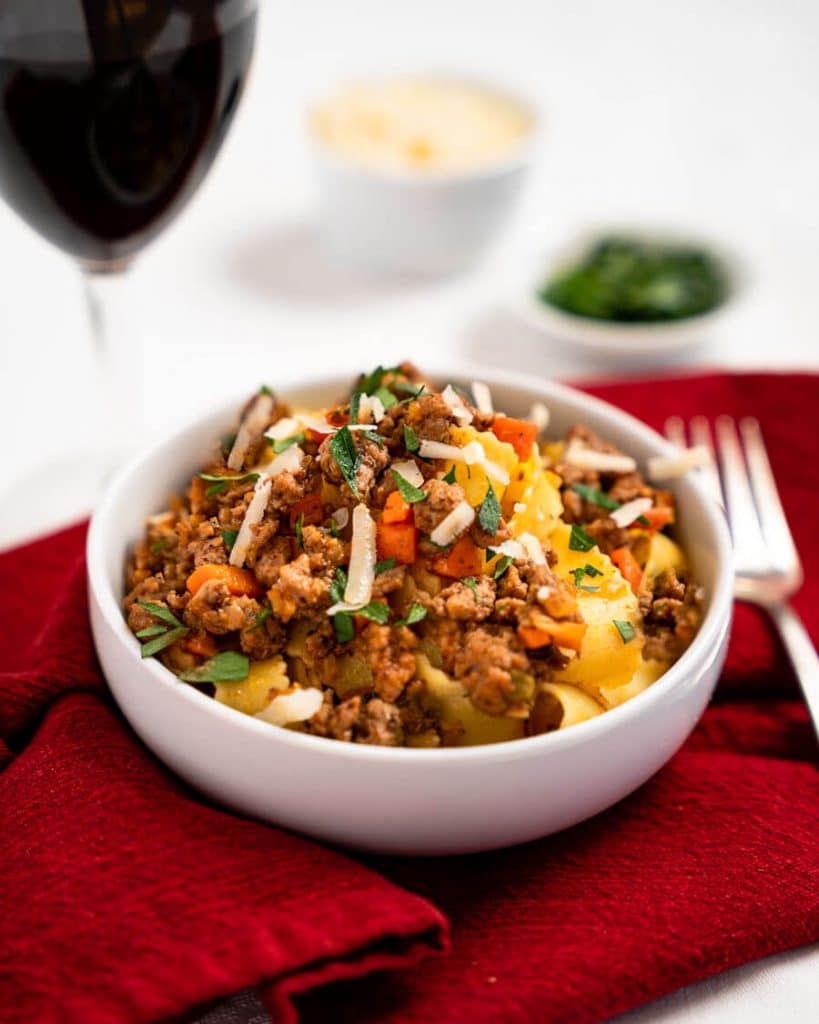 A bowl filled with ground lamb bolognese meat filling and pappardelle pasta garnished with parsley and parmesan.