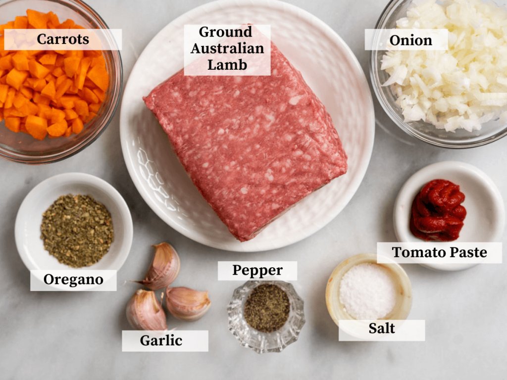 Top down photo of meat sauce ingredients including ground lamb, onion, carrots, tomato paste, garlic, Italian seasoning, salt, and pepper.