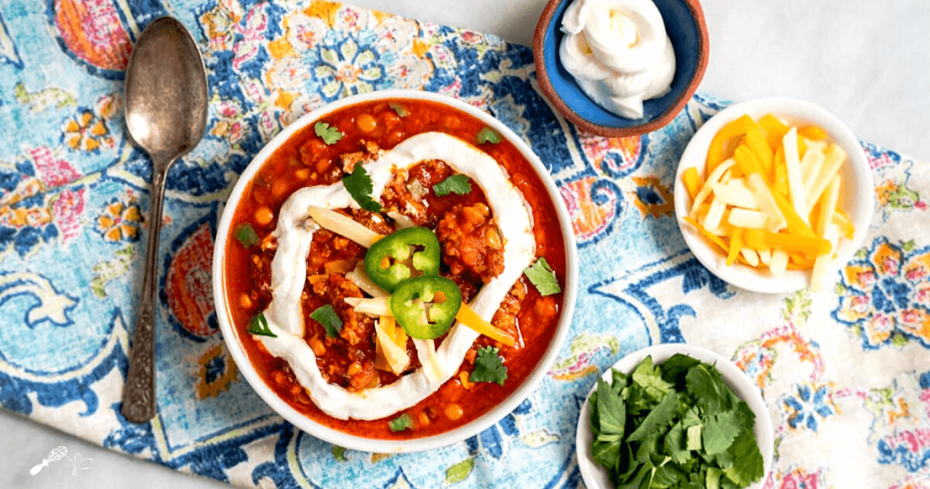 Top down photo of a bowl of lentil chili garnished with sour cream, sliced jalapenos, cilantro and cheese.