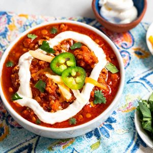 Close-up top down photo of a bowl of lentil chili garnished with sour cream, sliced jalapenos, cilantro and cheese.