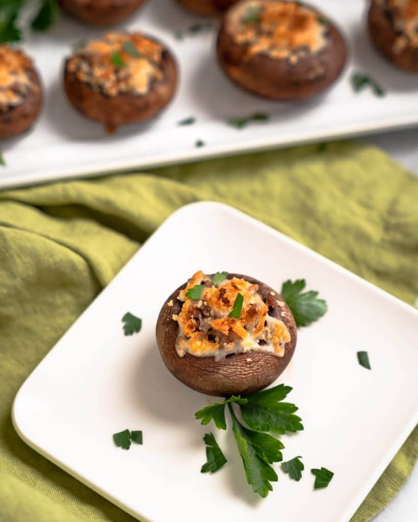 A mushroom stuffed with meat and cheese on a white plate with a tray of mushrooms in the background.