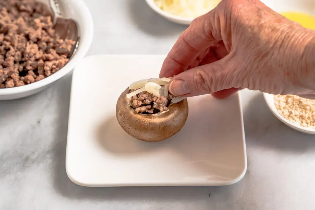 Shredded cheese being applied over meat filling in a mushroom cap.