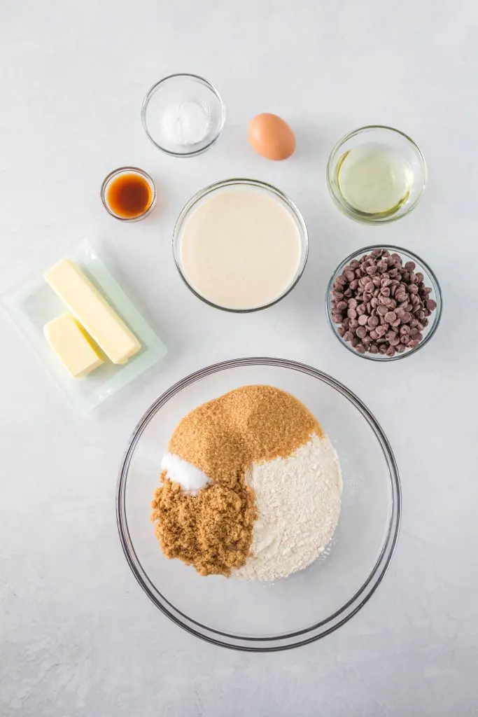 top down view of ingredients used to make sourdough chocolate chip cookies including sourdough starter, flour, sugar, vanilla, butter, baking soda, oil, egg, and salt and chocolate chips