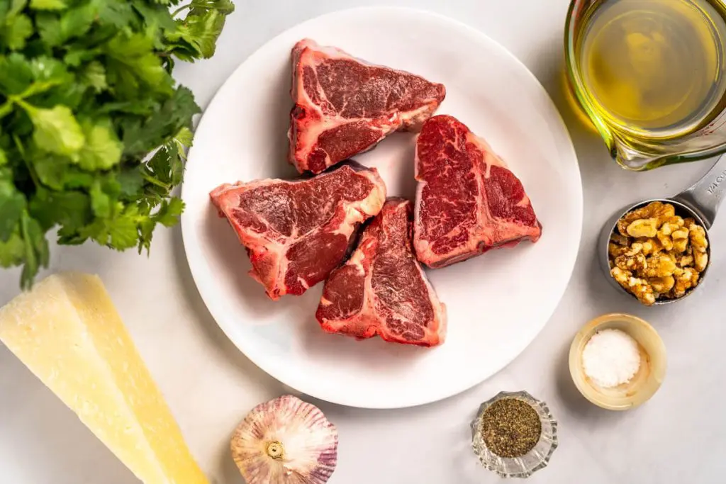 Top view of ingredients used to make grilled lamb loin chops including Lamb chops, Cilantro, Olive oil, Garlic, Walnuts, Salt, Pepper, and Romano Cheese.