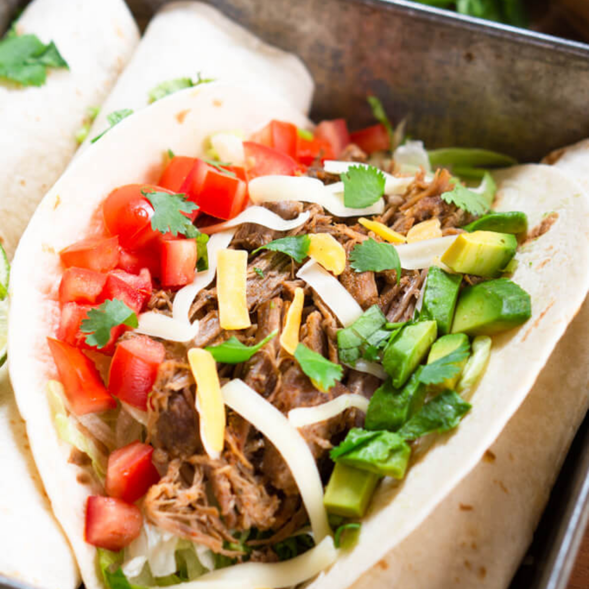 Angled view of a soft shell taco filled with shredded pork taco meat and garnished with diced tomatoes, avocados, and shredded cheese. Hostess At Heart