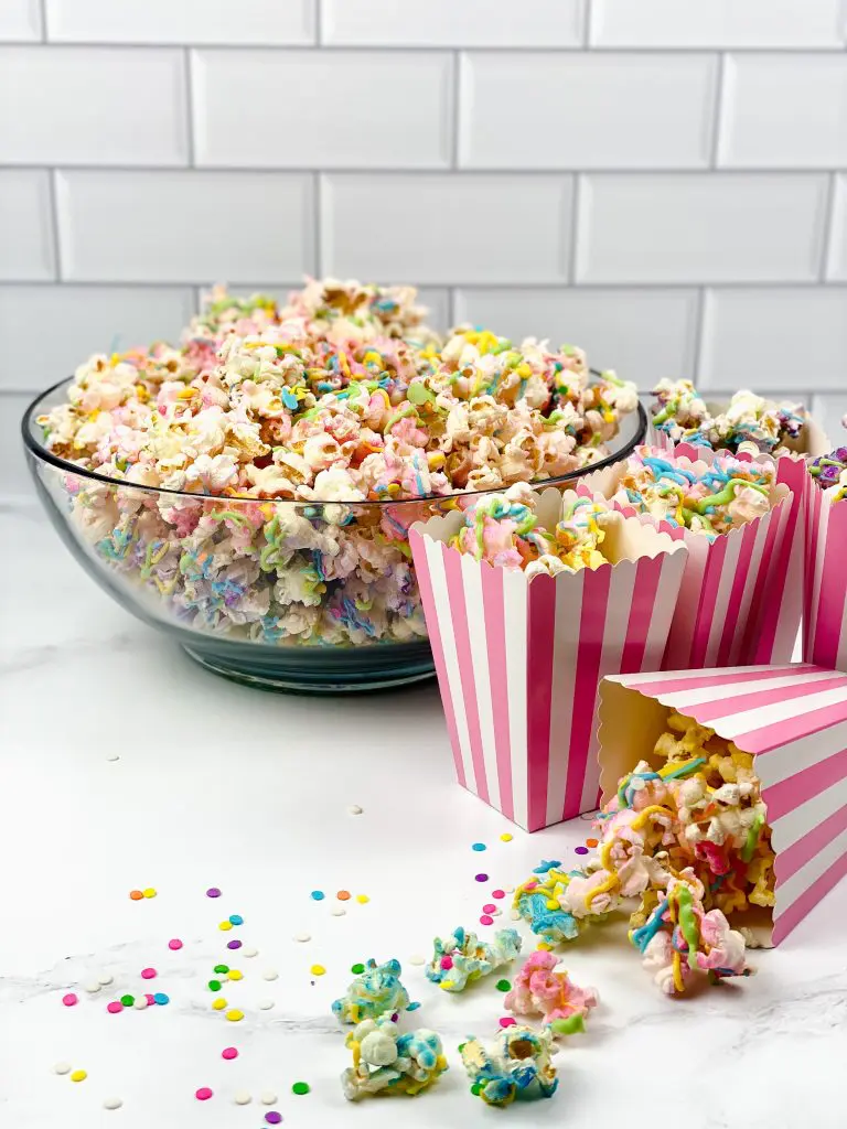 unicorn popcorn in a bowl on the counter with individual servings in pink and white striped single serving popcorn bags in front of a white tiled wall.