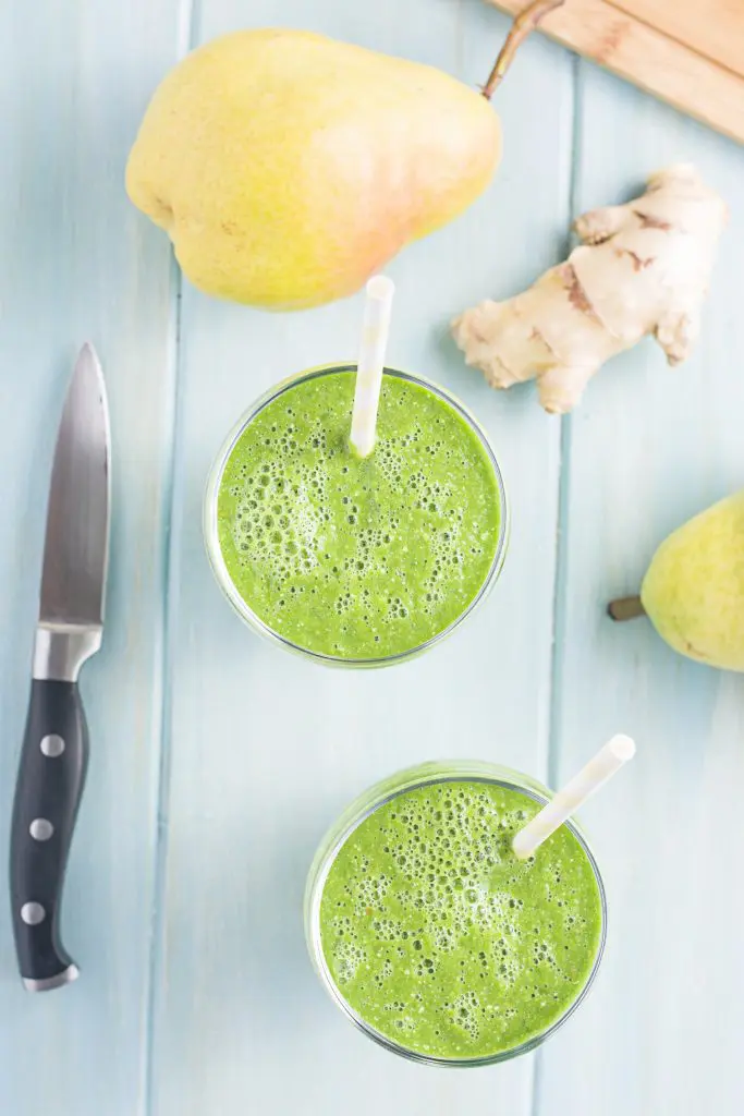 Green smoothie in a cup with ingredients around it. Shown are ginger and pears with a knife.