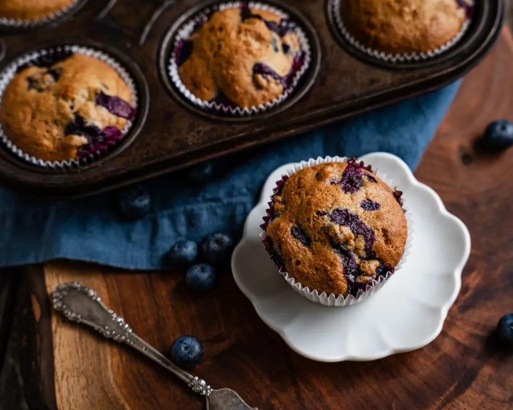 Top view of a blueberry banana muffin sitting on a small plate with the muffin pan filled with muffins behind it.