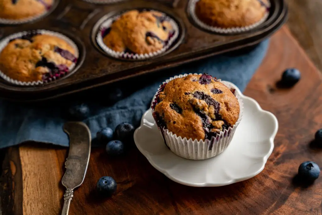 3/4 view of a blueberry banana muffin sitting on a white plate with the filled muffin tin behind it.