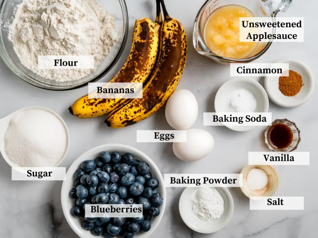 Top down view of the ingredients in banana blueberry muffins including flour, sugar, blueberries, bananas, eggs, cinnamon, vanilla, salt, baking powder and soda, cinnamon and applesauce