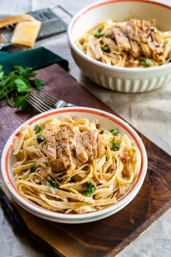 3/4 angle of a bowl of cooked fettuccine pasta topped with slices of grilled chicken and dusted with grated cheese.