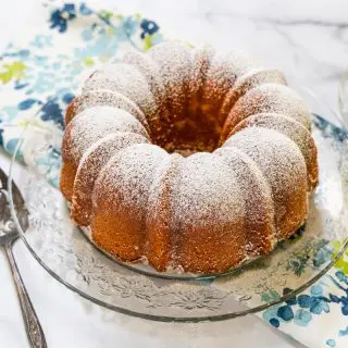 3/4 view of a powder sugar dusted bundt cake sitting on a cake plate over a multi-colored napkin.