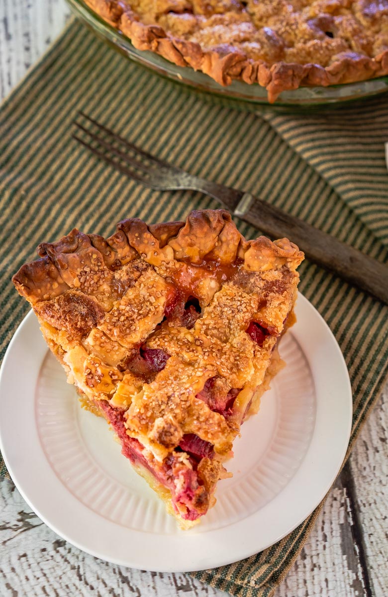 Top down view of a slice of rhubarb custard pie with a lattice top sitting on a plate with the whole pie in the background.