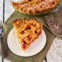 Top down view of a slice of rhubarb custard pie with a lattice top sitting on a plate with the whole pie in the background.
