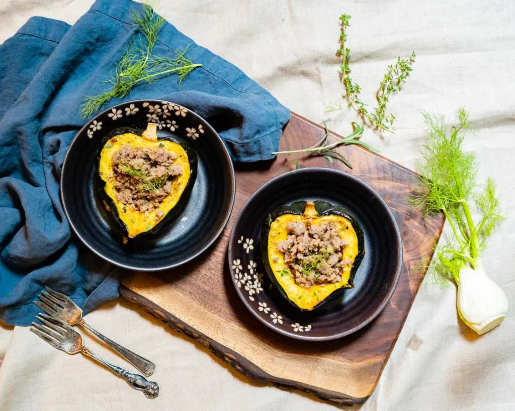 Two sausage-stuffed acorn squash halves, on a wooden board with herbs around. 