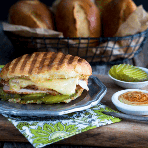 Oval Medianoche bread roll filled with dill pickle slices, ham, roasted pork, and Swiss cheese on a grey metal plate and then grilled. White dishes of a sriracha aioli and pickle slices sit next to a basket of Medianoche rolls.