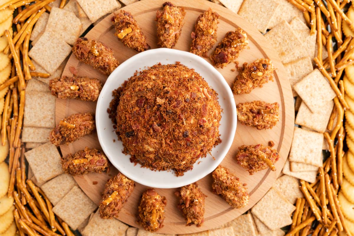 Top down view of a cheeseball and football cheeseballs rolled in bacon and served with crackers and pretzel sticks.