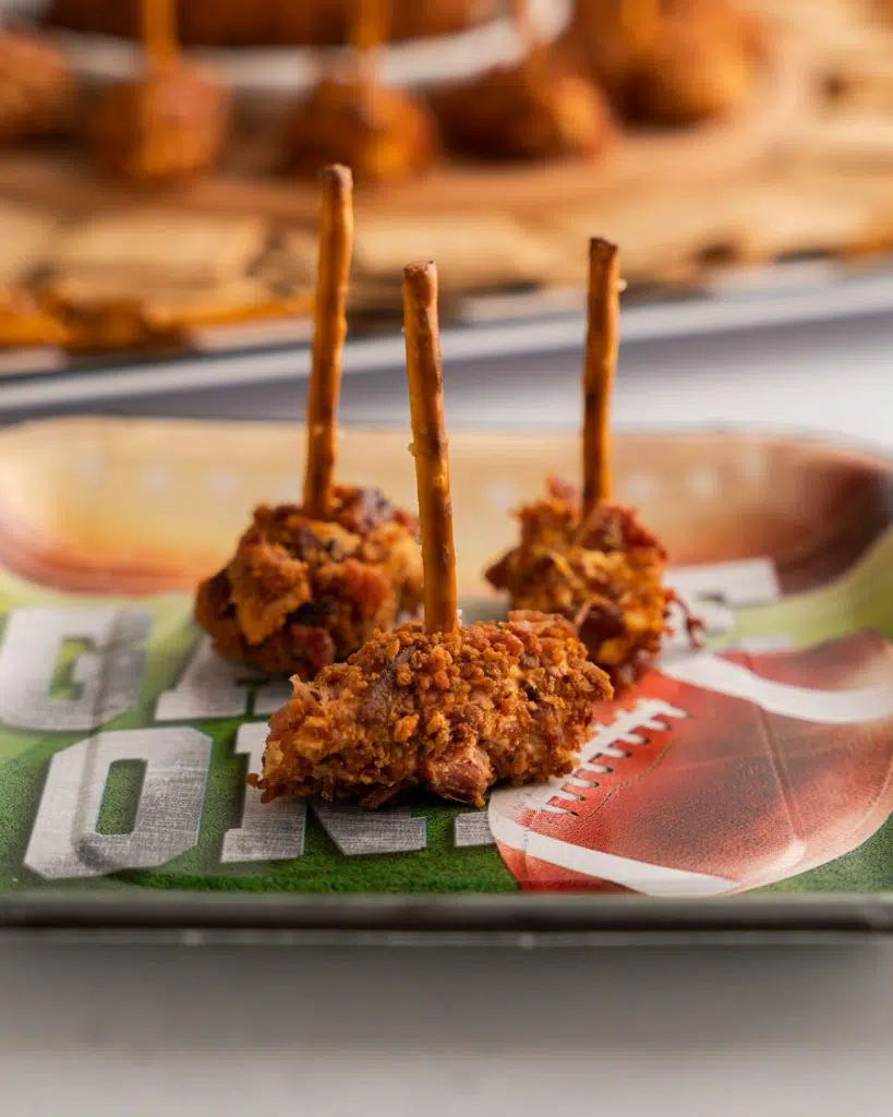 Close up table photo of three football shaped cheeseballs rolled in bacon bits and speared with pretzel sticks.