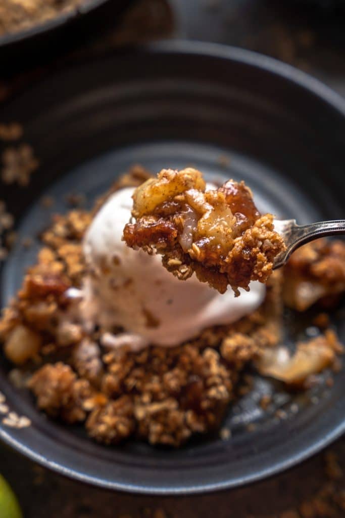 single bite of pear crisp in focus, with single serving of dish and ice cream shown behind the bite out of focus. 
