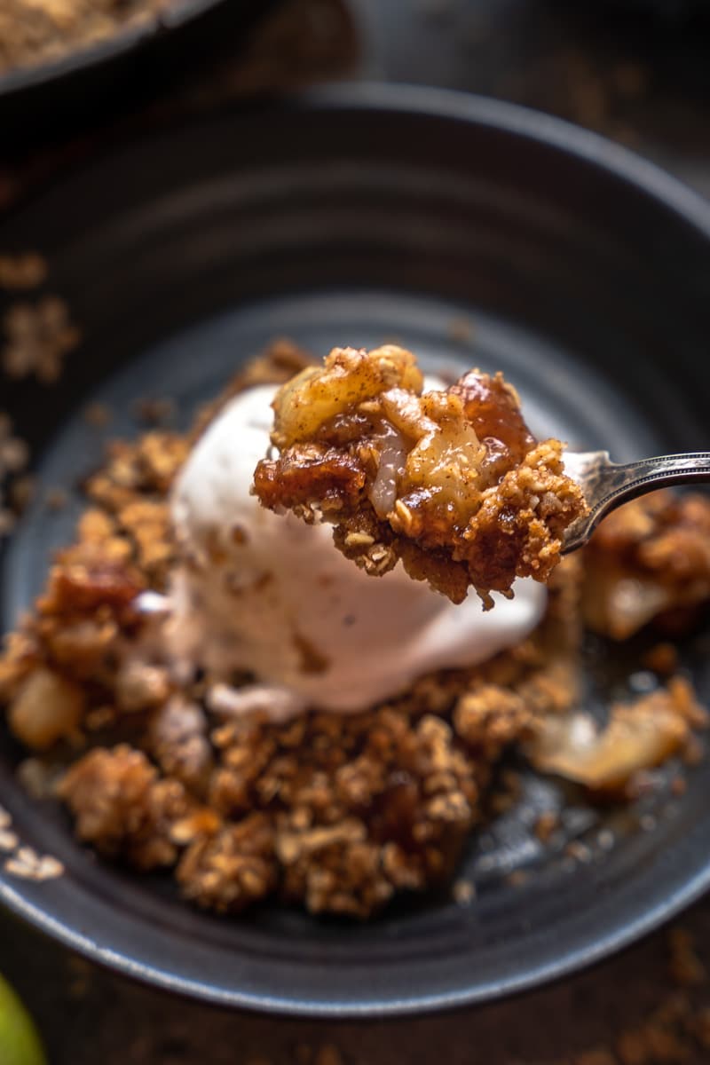 single bite of pear crisp in focus, with single serving of dish and ice cream shown behind the bite out of focus.