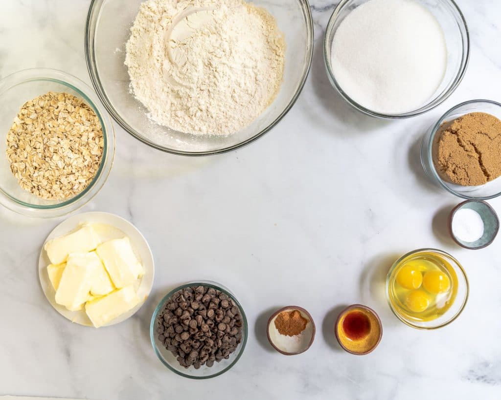 Top down view of chocolate chip oatmeal cookie ingredients including flour, sugar, butter, eggs, brown sugar, cinnamon, oats, and chocolate chips. Hostess At Heart