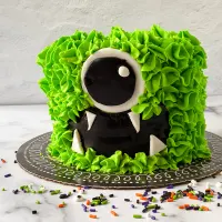 A cake decorated to look like a green monster with black eyes and fangs sprinkles scattered around it.