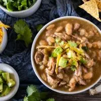 Top down view of a bowl of pork chili topped with avocado and cilantro with bowls of shredded cheese, cilantro, green onions, and tortilla chips off to the side.
