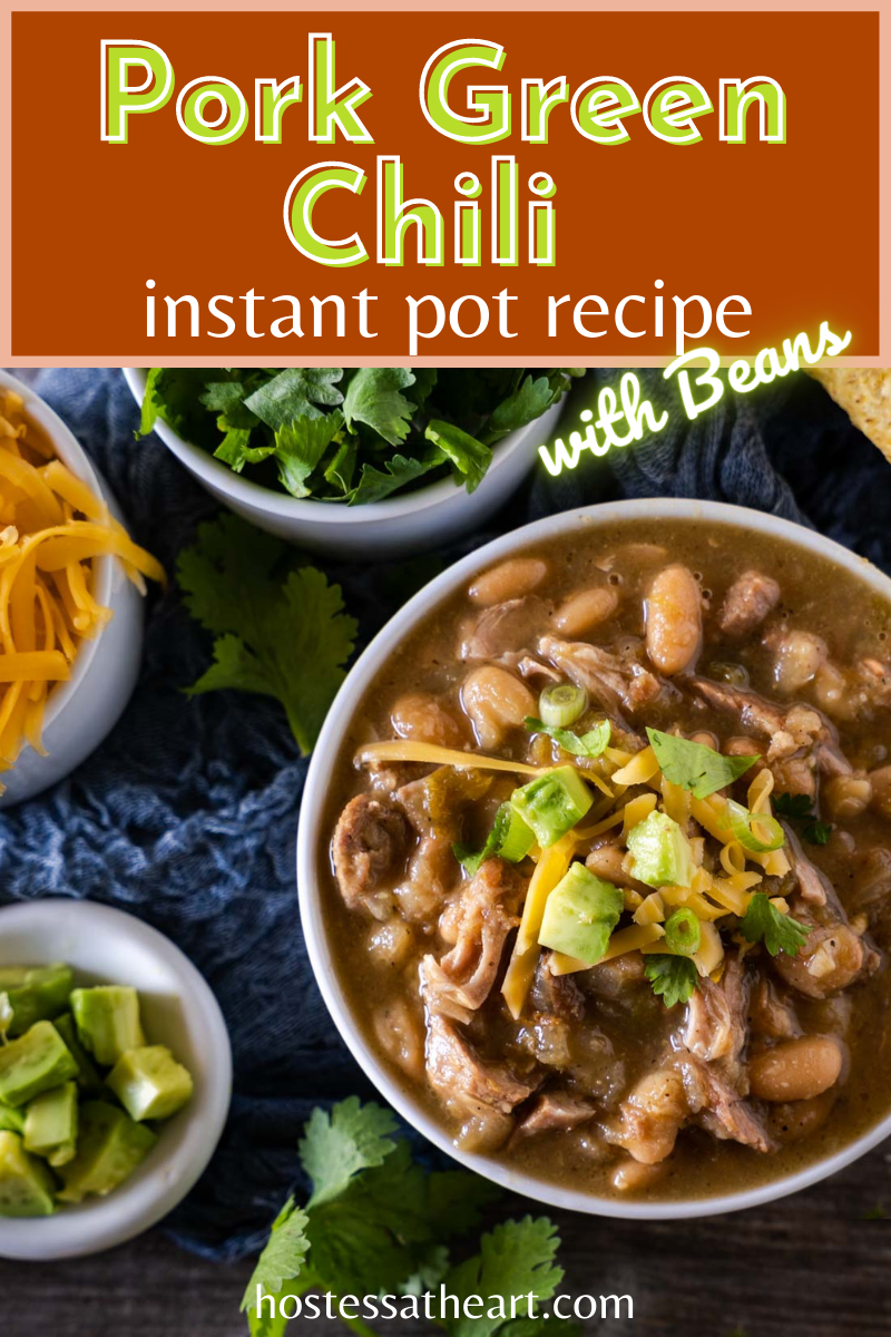 Top down view of a bowl of pork chili topped with avocado and cilantro with bowls of shredded cheese, cilantro, green onions, and tortilla chips off to the side.