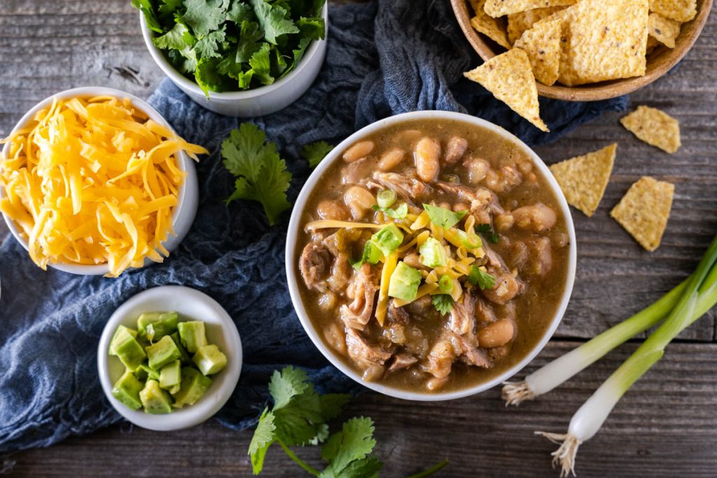 Top down view of a bowl of pork chili topped with avocado and cilantro with bowls of shredded cheese, cilantro, green onions, and tortilla chips off to the side.